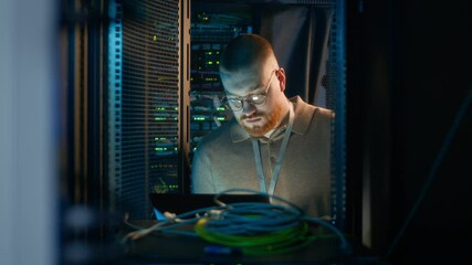 Medium shot of young Caucasian male specialist wearing glasses working on laptop in server room with hardware and big data - Powered by Adobe