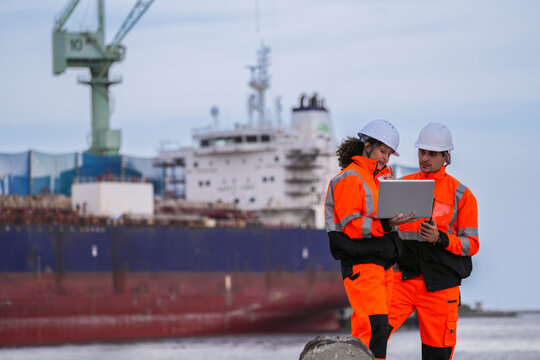 Two marine engineers wearing safety gear working on a dockside project using a laptop, with a large cargo ship and industrial crane in the background under cloudy sky.
