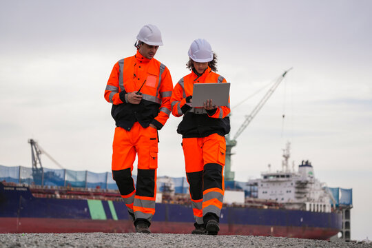Two marine engineers wearing safety gear working on a dockside project using a laptop, with a large cargo ship and industrial crane in the background under cloudy sky.