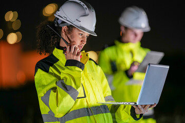 Two engineers wearing high-visibility suits review data on a laptop and tablet at night in front of...