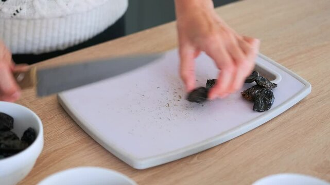 Close up of female hands cutting prunes with sharp knife on white cutting board on kitchen table. The process of preparing ingredients for a dish of porridge or smoothie.