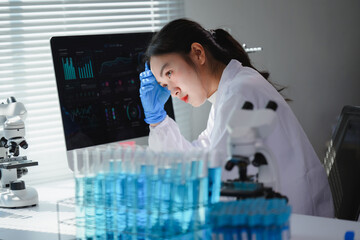 Asian female scientist looking concerned while analyzing data on computer in laboratory with test tubes and microscope