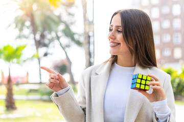 Young pretty woman holding a three dimensional puzzle cube at outdoors pointing to the side to present a product