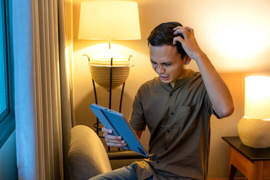 Frustrated man scratching head while looking at tablet in warm-lit living room - Powered by Adobe