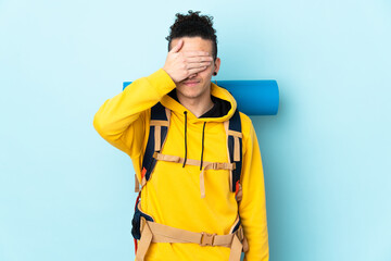 Young mountaineer man with a big backpack over isolated blue background covering eyes by hands