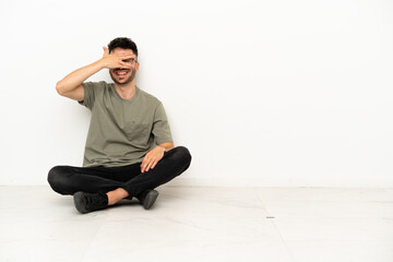 Young caucasian man sitting on the floor isolated on white background covering eyes by hands and smiling
