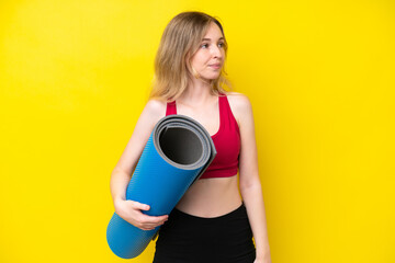 Young sport caucasian woman going to yoga classes while holding a mat isolated on yellow background...