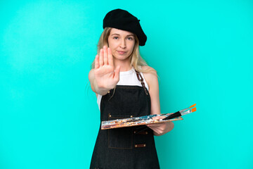 Young artist caucasian woman holding a palette isolated on blue background making stop gesture