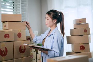 An Asian woman uses a tablet to manage online orders and prepare packages for shipping —...
