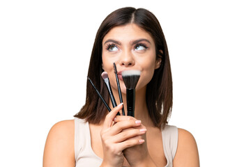 Young Russian woman with rainproof coat and umbrella over isolated chroma key background holding makeup brush and looking up
