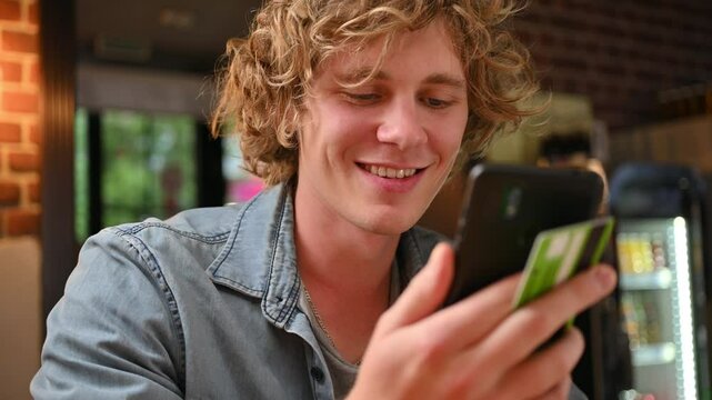 A young man calculates on a telephone calculator, smiles, talks to his interlocutor at the table and pays with a bank credit card in a cafe.