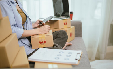 An Asian female entrepreneur prepares parcels and checks online orders from her home using her laptop and smartphone, reflecting the concept of online shopping and starting a SME business.