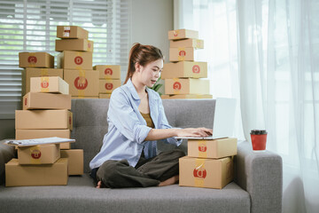 A young Asian female entrepreneur prepares packages and checks online orders from her home using a laptop, representing the concept of online shopping and a growing small SME startup