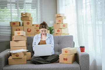 A young Asian female entrepreneur prepares packages and checks online orders from her home using a laptop, representing the concept of online shopping and a growing small SME startup