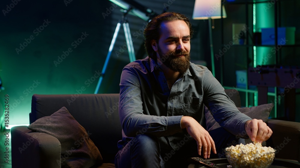 Poster man enjoying bowl of popcorn and smiling while watching cable programs on tv during leisure time at 