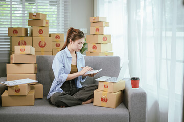 An Asian female entrepreneur prepares parcels and checks online orders from her home using her laptop and smartphone, reflecting the concept of online shopping and starting a SME business.