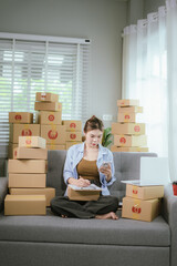 An Asian woman entrepreneur prepares parcels and manages her product orders via smartphone, ready to ship them to customers — reflecting the rise of online shopping and small business startups.
