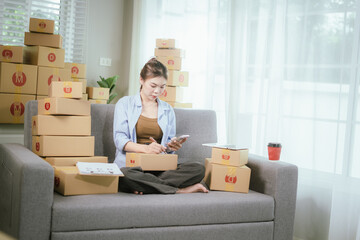 An Asian woman entrepreneur prepares parcels and manages her product orders via smartphone, ready to ship them to customers — reflecting the rise of online shopping and small business startups.