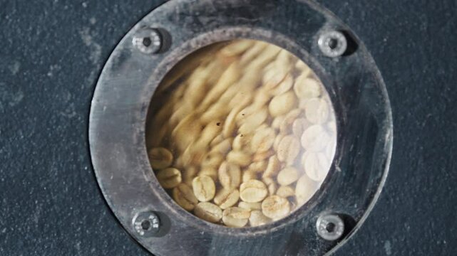 Green coffee beans tumbling inside industrial roasting machine. Rotating through circular metal window during precise manufacturing process. Transforming raw ingredients into aromatic roasted product