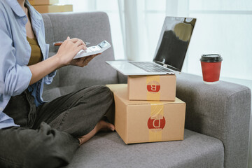 A young Asian female entrepreneur prepares packages and checks online orders from her home using a laptop, representing the concept of online shopping and a growing small SME startup