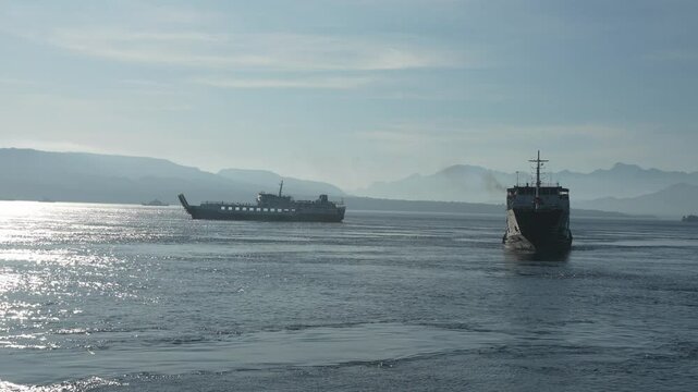 Two ferries cross Bali Strait carrying passengers to Gilimanuk Port. Shot from onboard one ferry approaching the harbor, capturing the spirit of travel and island connection.