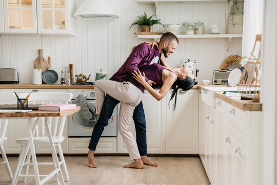 A couple dances intimately in the kitchen, sharing laughter amidst the day-to-day, framed by warm wood tones and comforting kitchenware.