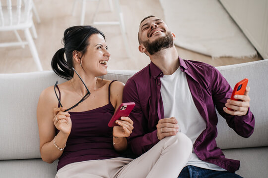 A jubilant couple sits on a couch laughing together as they browse their smartphones, highlighting a moment of shared humor and connection.