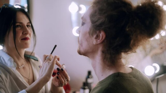 A female makeup artist carefully applies shimmering glitter to a male model&rsquo;s eyelids using her finger, creating a dazzling makeup look.