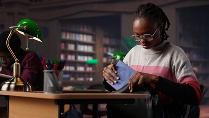 African american student comparing class notes with data from the library collection database, using laptop to focus on a literature assignment. Young pupil working for her diploma. Camera A.