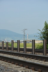 Fototapeta premium Train tracks recede into the distance with a row of short, rusty poles, under a clear sky with an industrial building and mountains beyond.