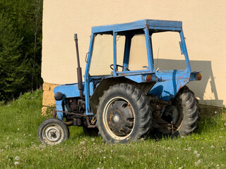 Obraz premium Old blue tractor parked near a wall on grass. Used only rarely on a farm. Rusty and aged, but still functional. Rural outdoor photo in daylight.
