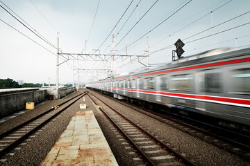 A blur of a train passes a station platform with tracks and railway signals under a cloudy sky,...