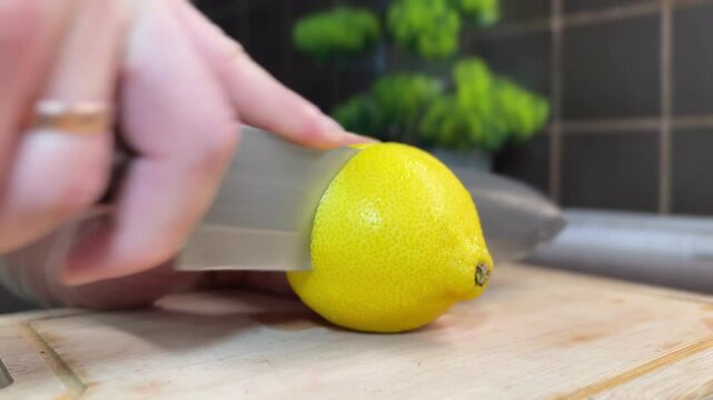 Close-up of slicing a fresh, ripe lemon with a large knife on a wooden cutting board at home. Juicy lemon preparation in a cozy kitchen environment.