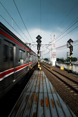 A blur of a train passes a station platform with tracks and railway signals under a cloudy sky,...