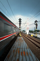 A blur of a train passes a station platform with tracks and railway signals under a cloudy sky,...