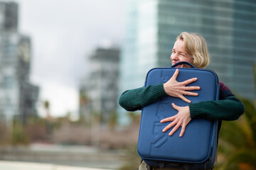 Woman with blond hair playfully hugging suitcase outdoors in cityscape