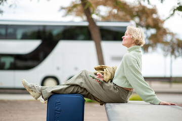 Relaxed woman with short blond hair waiting at bus station with legs on suitcase