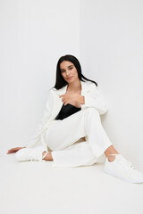 Woman wearing white suit and sneakers sitting relaxed on white floor by white wall, confident young female with long dark hair in casual stylish outfit in minimalistic setting.