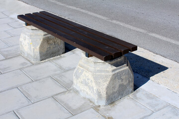 Simple park bench with dark wooden slats and sturdy concrete supports stands on a paved sidewalk by the road, casting a shadow in bright sunlight, offering a resting spot in an urban setting