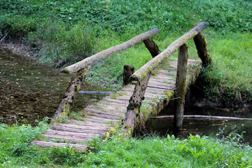 Narrow rustic footbridge made of rough wooden planks and logs with simple railings crosses a...