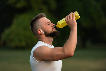 A young fit man with closed eyes listening to music on wireless headphones and drinking water or...