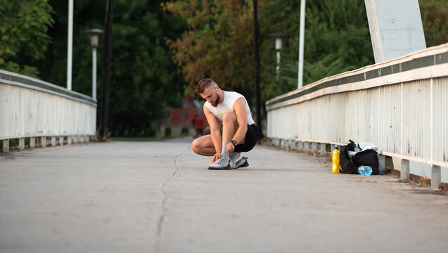 Young athletic man tying shoelaces before a workout on a city bridge at sunrise. Outdoor fitness preparation, active lifestyle concept.