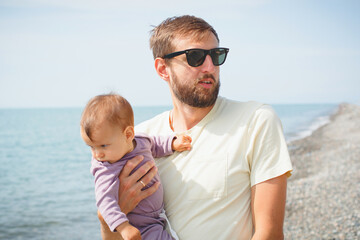 Young bearded father in sunglasses holding his baby dressed in purple near the seaside on a pebble...