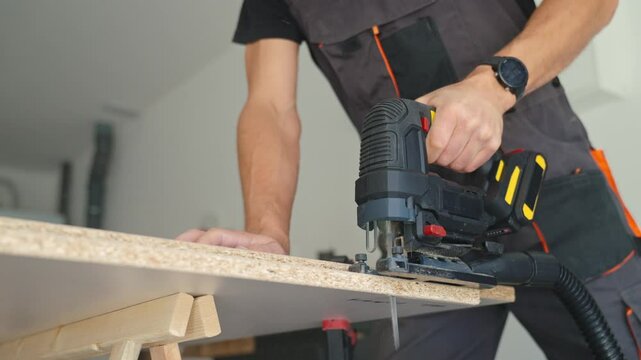 Man in workwear using jigsaw to cut chipboard panel in modern home workshop. Concept of DIY renovation, craftsmanship and home improvement process.
