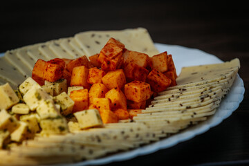 Close-up of a cheese platter featuring spiced and herbed cheese cubes, and thinly sliced cheese arranged on a white plate over a dark surface.
