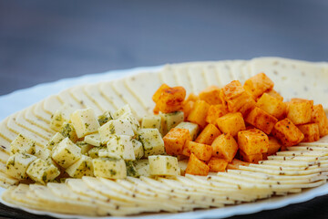 Close-up of a cheese platter featuring spiced and herbed cheese cubes, and thinly sliced cheese arranged on a white plate over a dark surface.