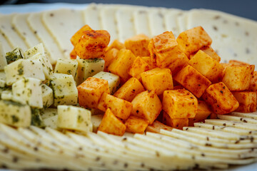 Close-up of a cheese platter featuring spiced and herbed cheese cubes, and thinly sliced cheese arranged on a white plate over a dark surface.