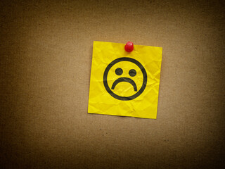 A yellow paper note with a sad face on it pinned to cardboard. Depression concept. Close up.
