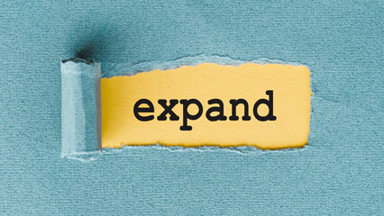 A close-up shot of a torn piece of blue paper revealing a yellow piece of paper with the word expand written in black lettering.
