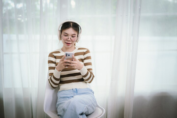 An Asian woman wearing headphones and glasses is casually using a smartphone in front of a window.